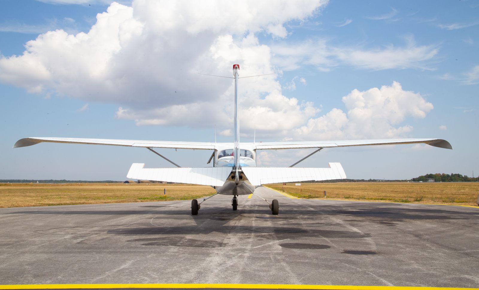 A white high-wing single-engine aircraft seen from behind on a runway under big summer clouds.