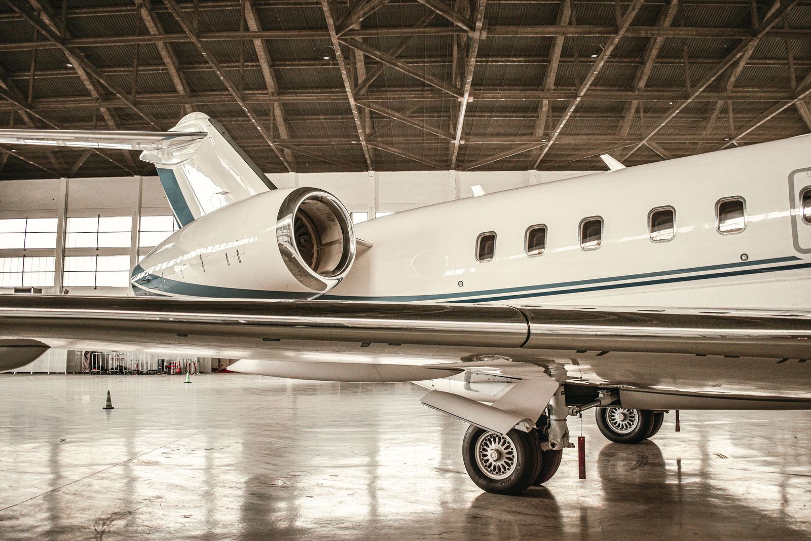 A white business jet with blue stripes parked in a hangar, its wing and engine reflected in the polished floor.