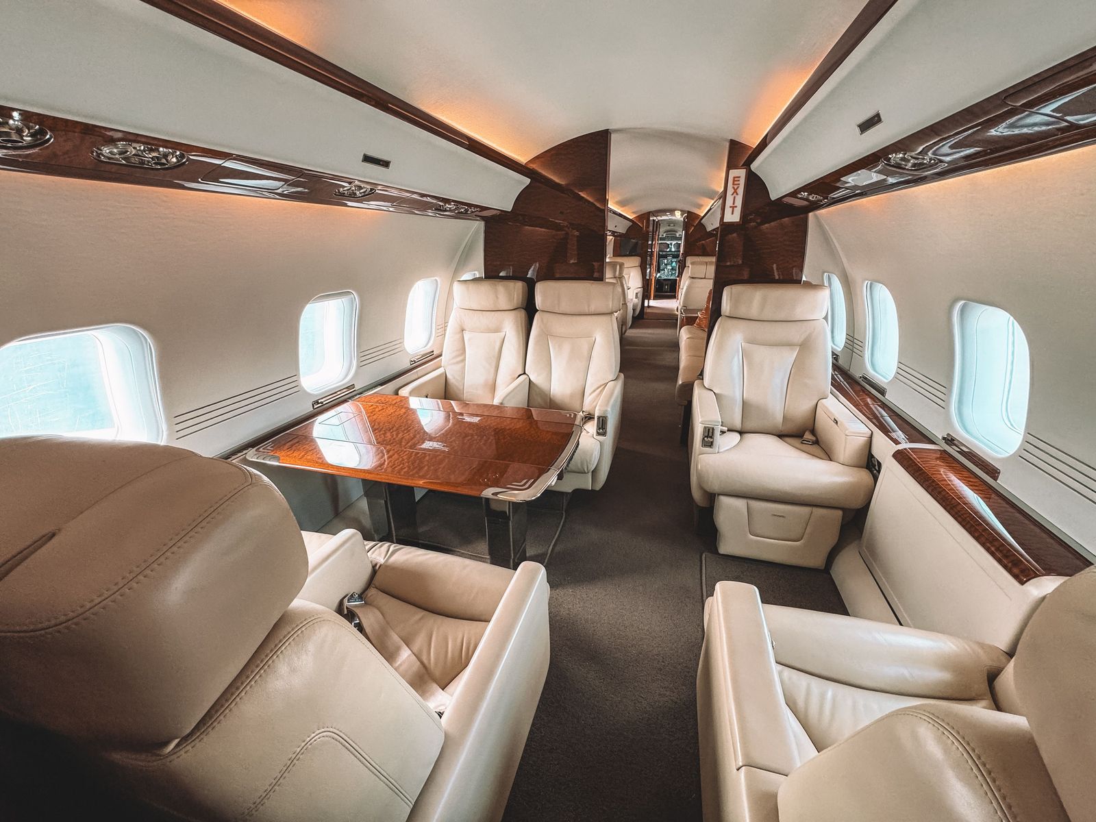 A tidy business jet cabin with cream leather club seats, a polished wood table and oval windows.