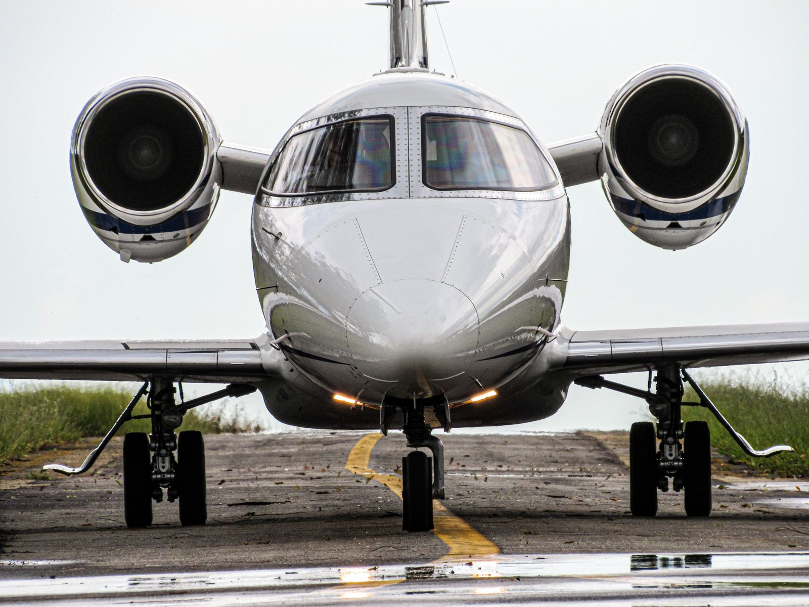 Head-on view of a glossy white business jet on a wet taxiway, its nose and engine intakes reflecting the sky.